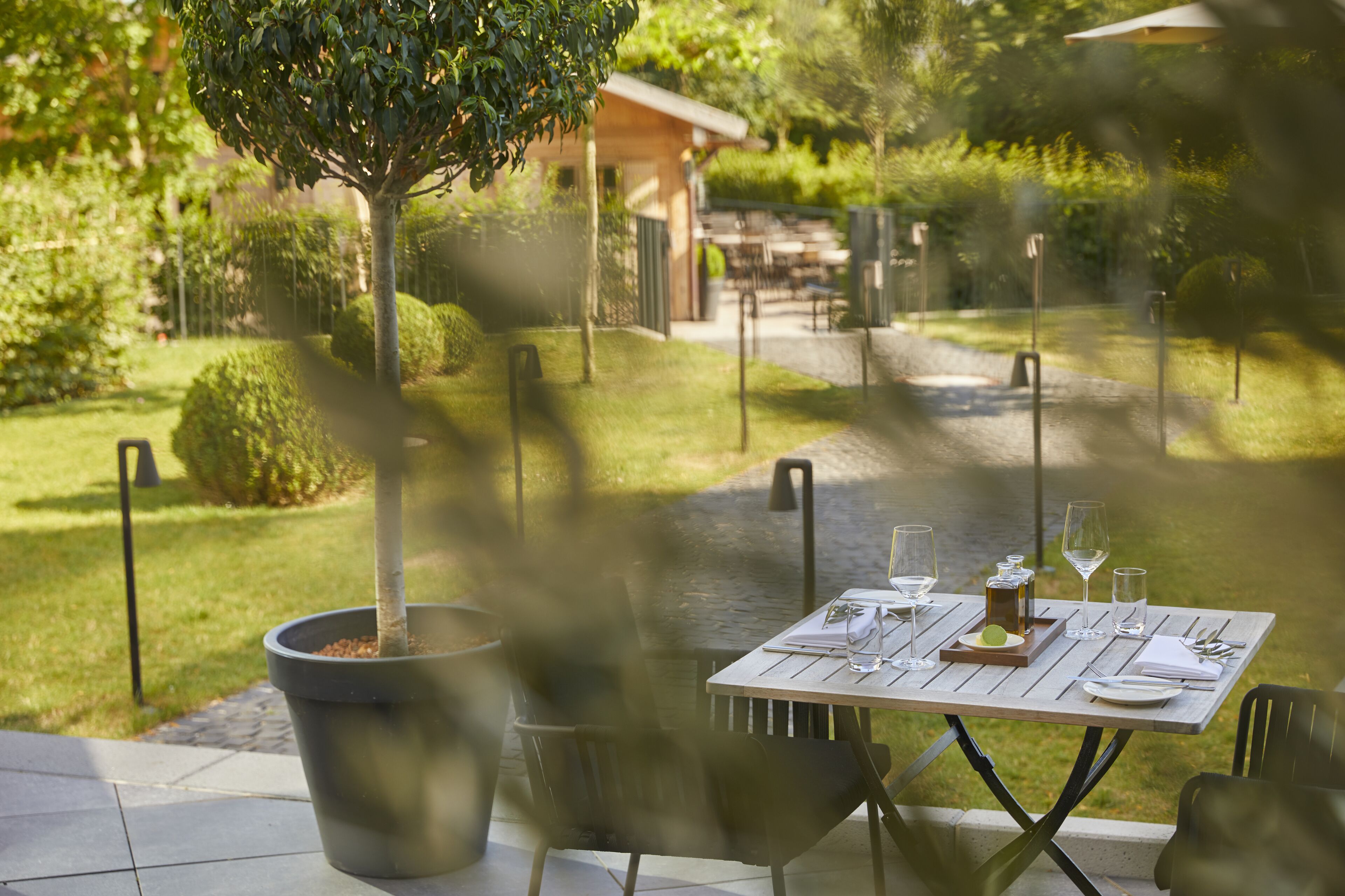 An outdoor dining area with a table set for two, surrounded by potted plants and a view of a garden and building in the background.