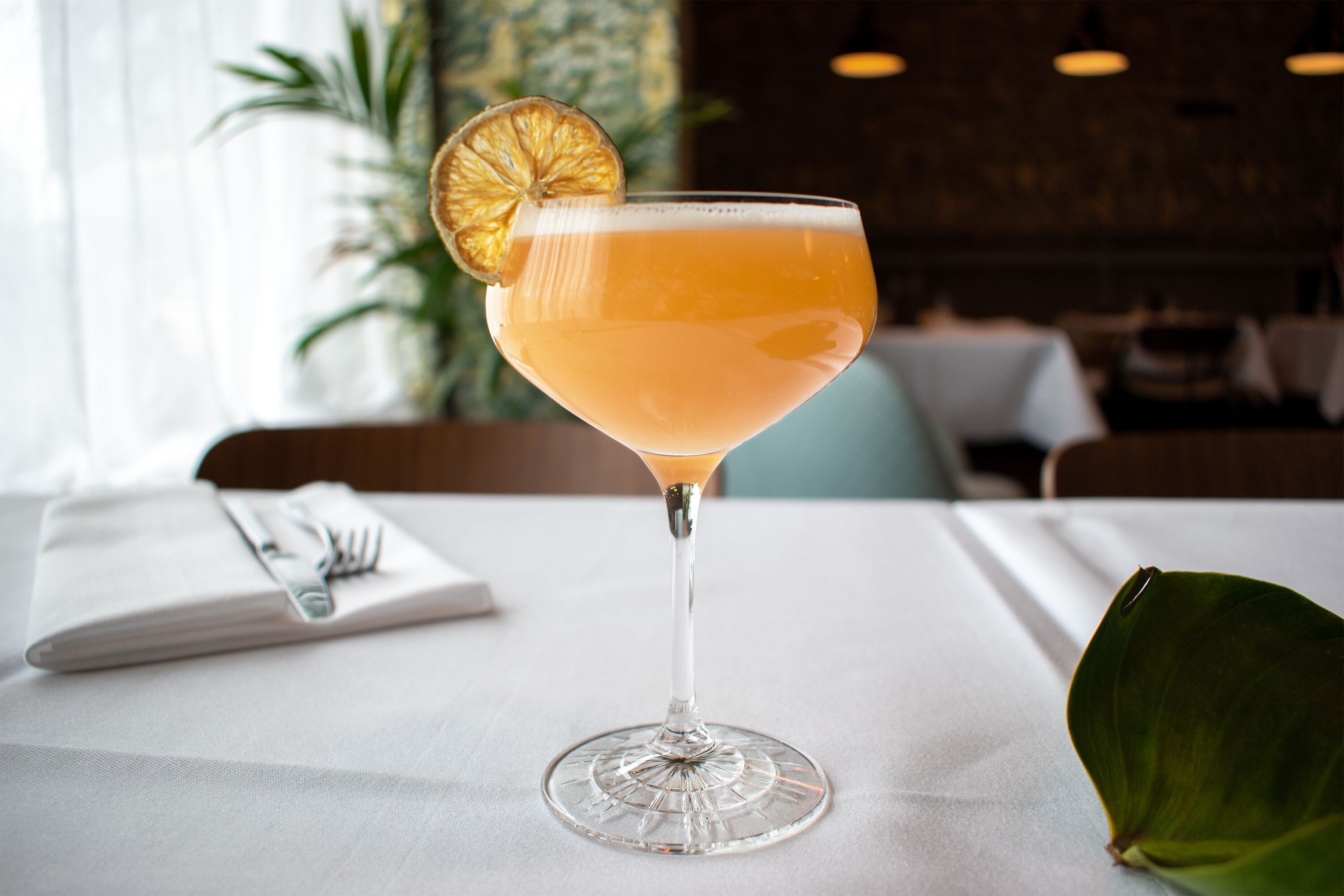 A cocktail in a martini glass with a lemon garnish on a table in a restaurant.