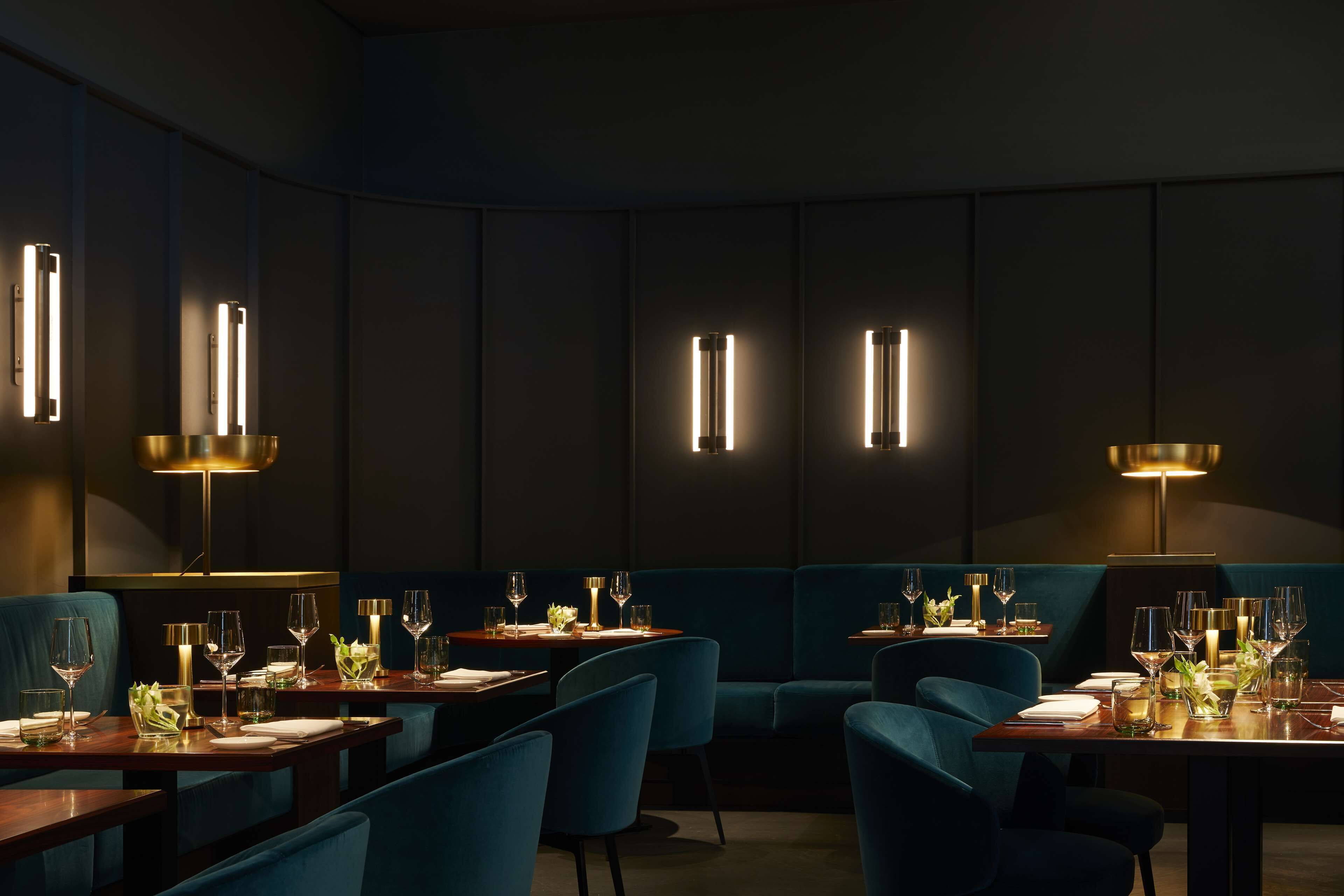 Ein Restaurant in gedämpftem Licht mit dunklen Wänden, blau bezogenen Sitzmöglichkeiten und Tischen, die mit Weingläsern und Kerzen gedeckt sind.
