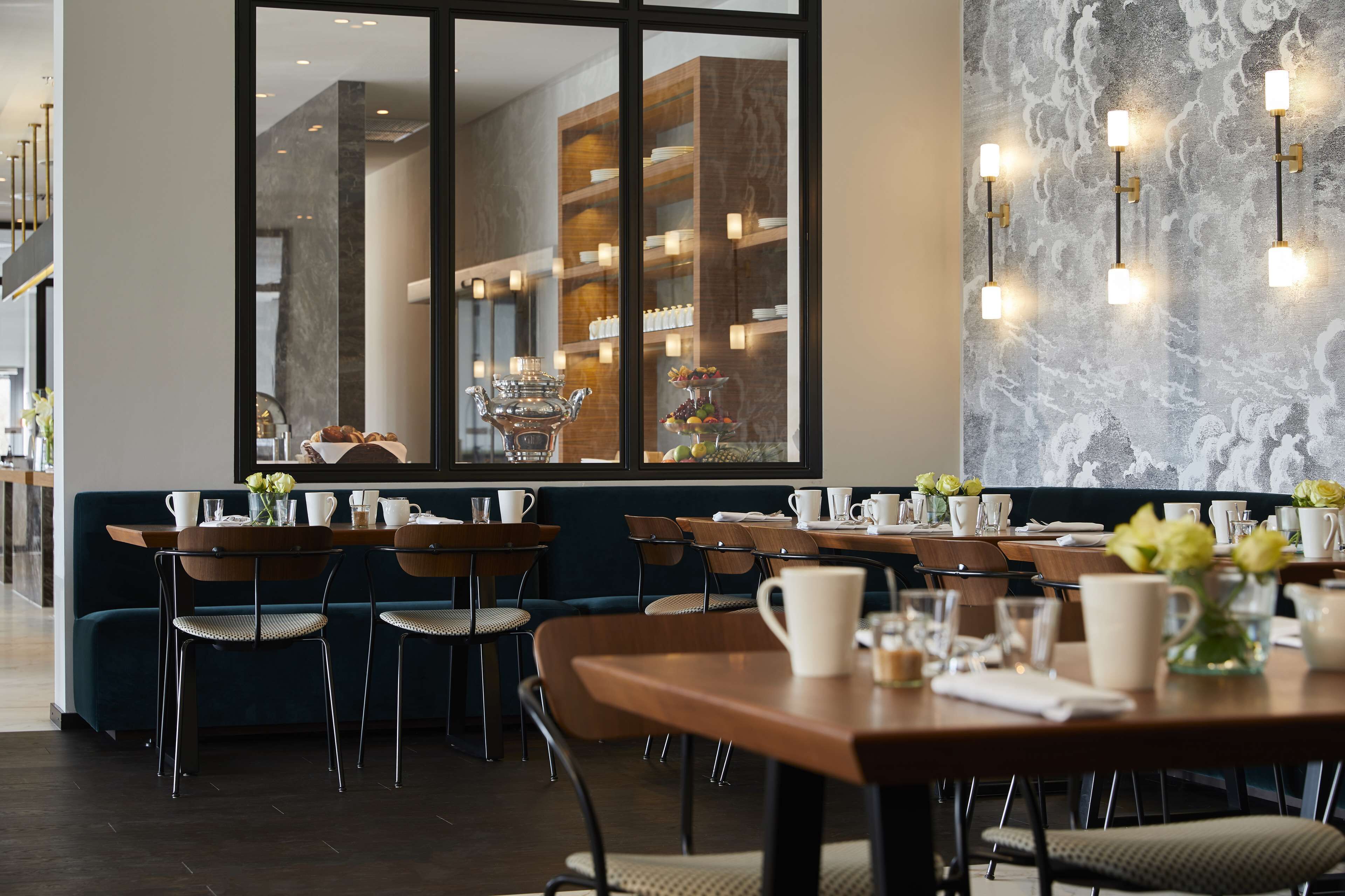 Un comedor moderno con mesas y sillas de madera, un ventanal con vista a la cocina y apliques decorativos en la pared.