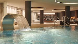 A modern indoor pool area with a waterfall feature, lounge seating, and a staircase leading to a higher level. - Infinity Hotel & Conference Resort Munich (Unterschleissheim)