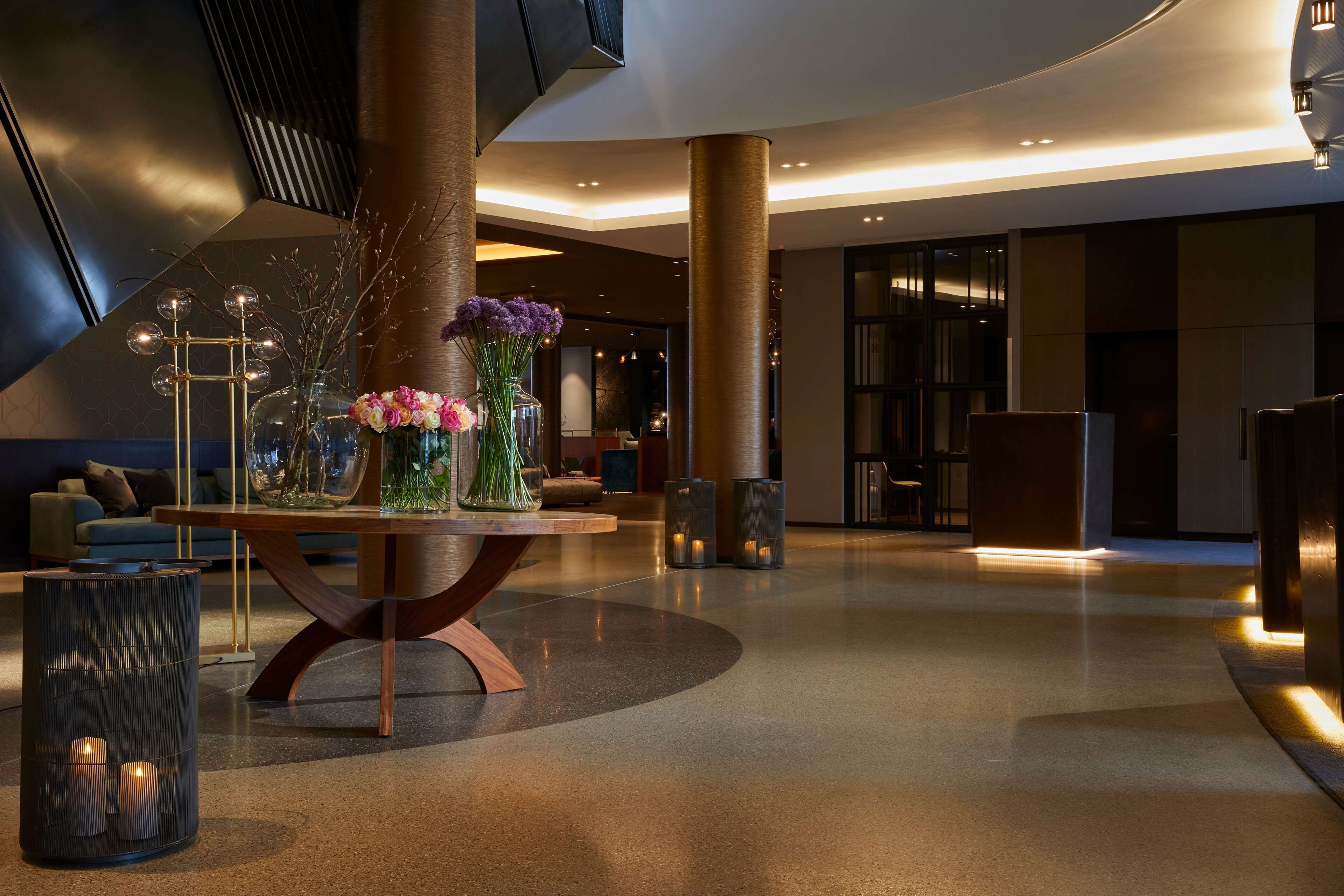 Eine moderne Lobby mit einem zentralen Tisch, der Vasen und Kerzen präsentiert, umgeben von Sitzgelegenheiten und einer großen Säule.