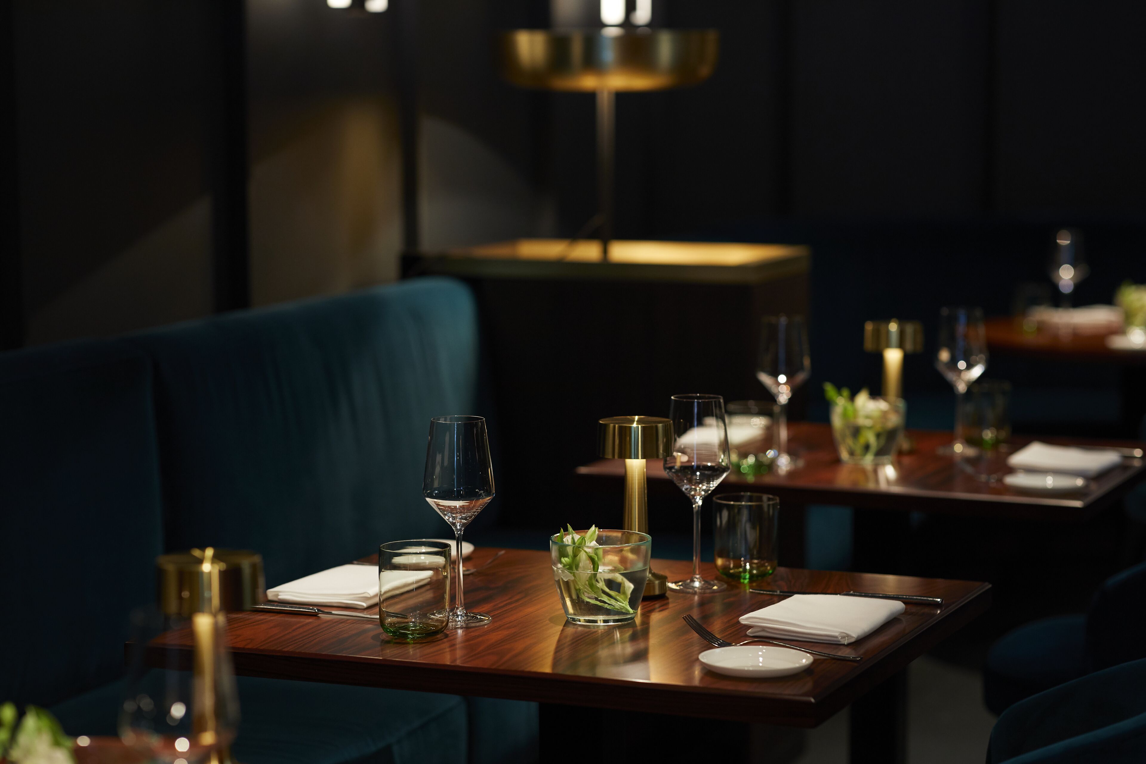 A dimly lit restaurant with tables set for dining, featuring elegant table settings and a modern chandelier.