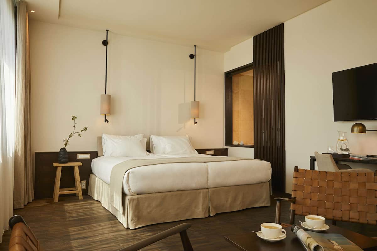 Une chambre d’hôtel avec un grand lit, deux tables de chevet, un téléviseur à écran plat fixé au mur, une chaise en bois et une petite table avec une tasse et une soucoupe.