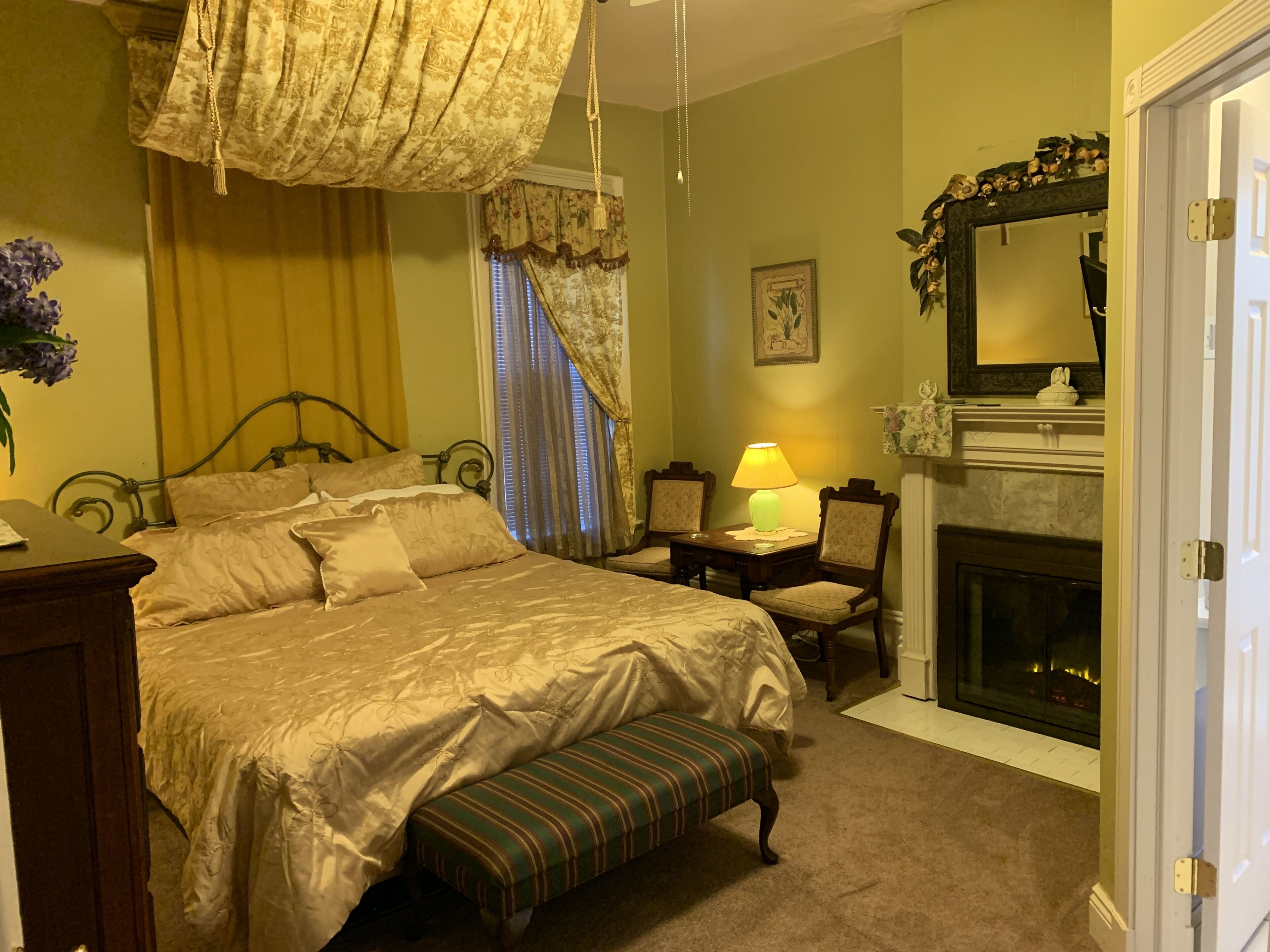 A bedroom with a large bed, a bench, a desk with a lamp, a fireplace, and a mirror.