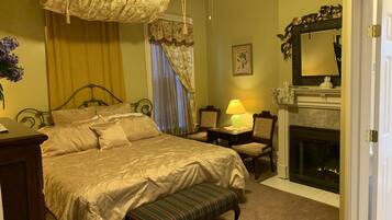 A bedroom with a large bed, a bench, a desk with a lamp, a fireplace, and a mirror.