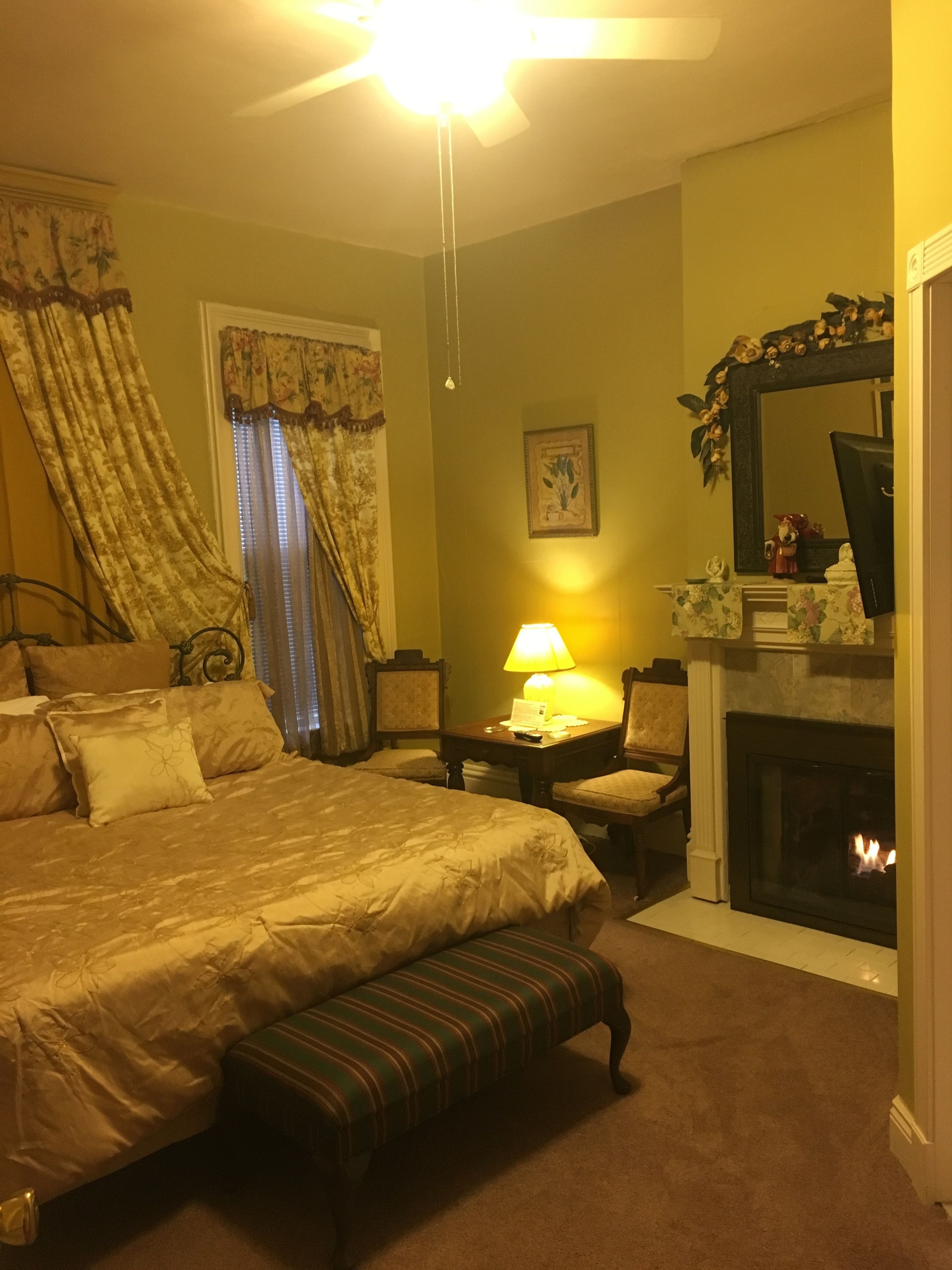 Un dormitorio de hotel con una cama grande, una mesita de noche con lámpara, una silla, una chimenea y un espejo sobre la chimenea.