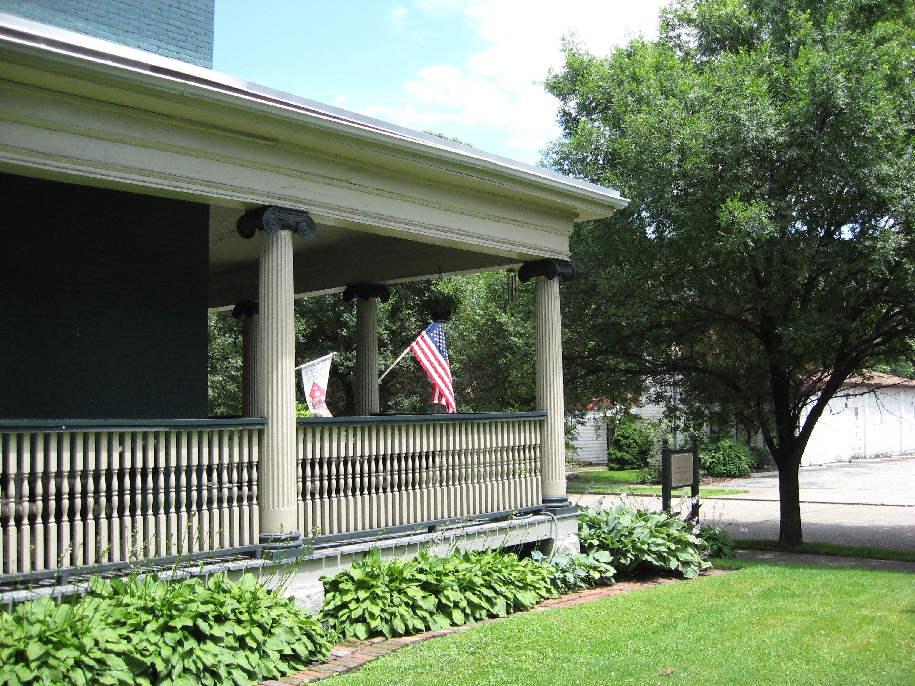 A building with a covered porch supported by columns, a flag, and greenery.