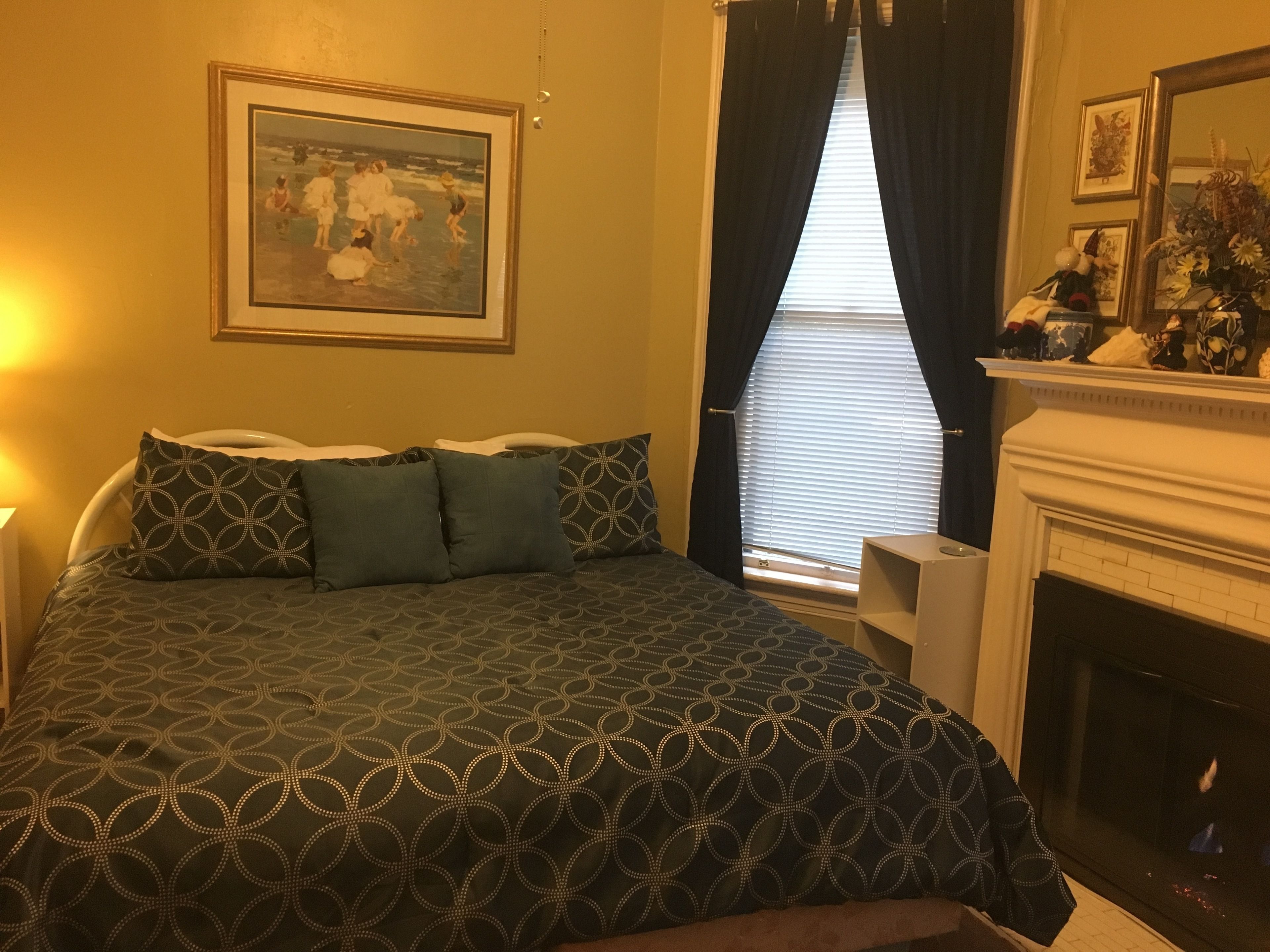 A bedroom with a bed, a painting on the wall, a window with curtains, and a fireplace.