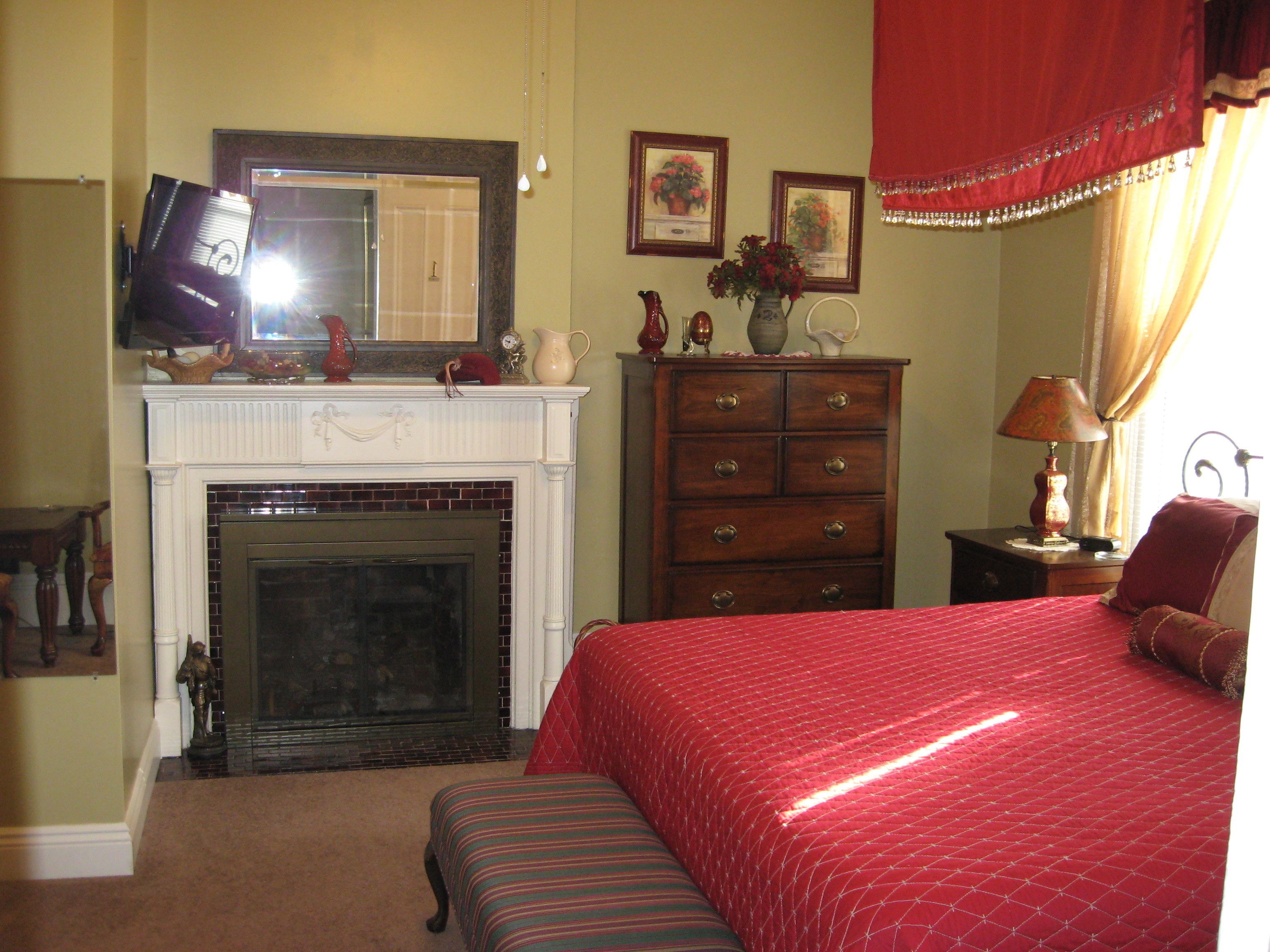 A bedroom with a fireplace, a bed with a red quilt, a wooden dresser, and a television on a stand.