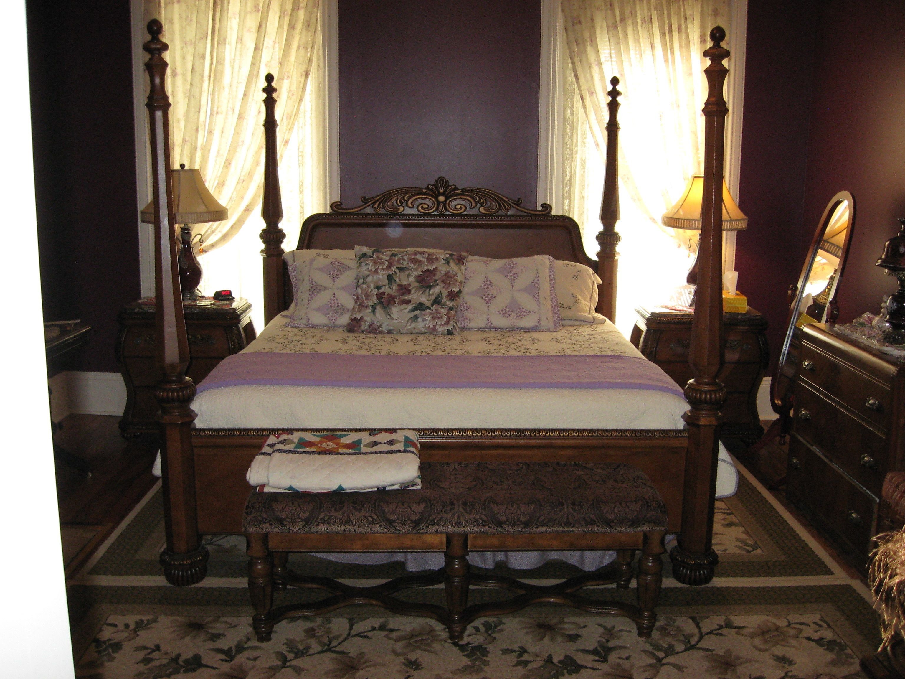 Uma cama de dossel com colcha e almofadas florais, ladeada por duas mesas de cabeceira com abajures, em um quarto com carpete estampado e espelho na parede.