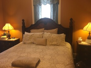 Un lit bien fait, recouvert d’une couvre-lit à motifs floraux, deux lampes de chevet, une table de chevet avec une horloge et un vase de fleurs, et une fenêtre avec des rideaux.