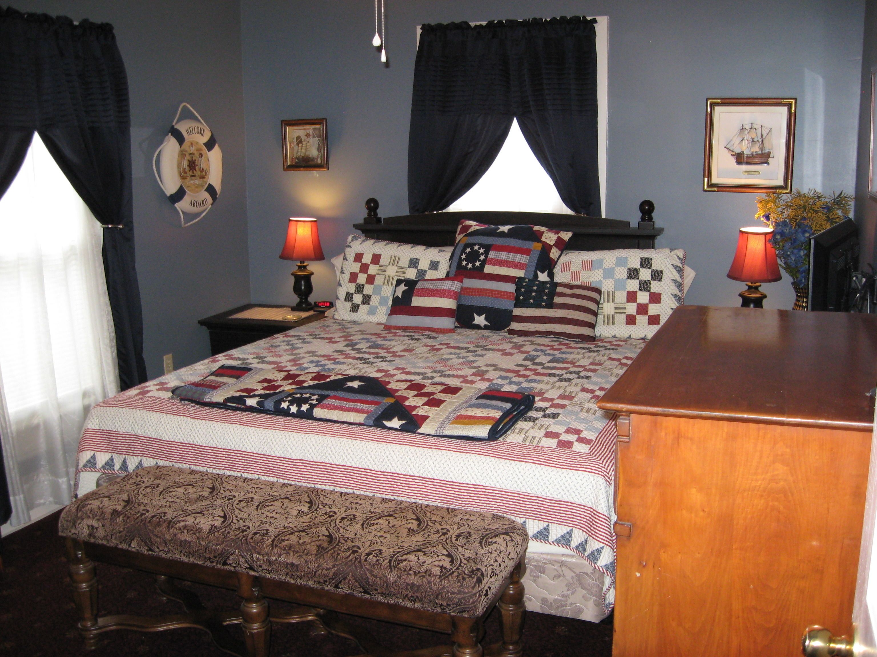 A neatly made bed with a quilt featuring a patriotic design, a wooden bench at the foot of the bed, two bedside lamps, and a framed picture on the wall.