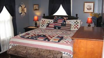 A neatly made bed with a quilt featuring a patriotic design, a wooden bench at the foot of the bed, two bedside lamps, and a framed picture on the wall.