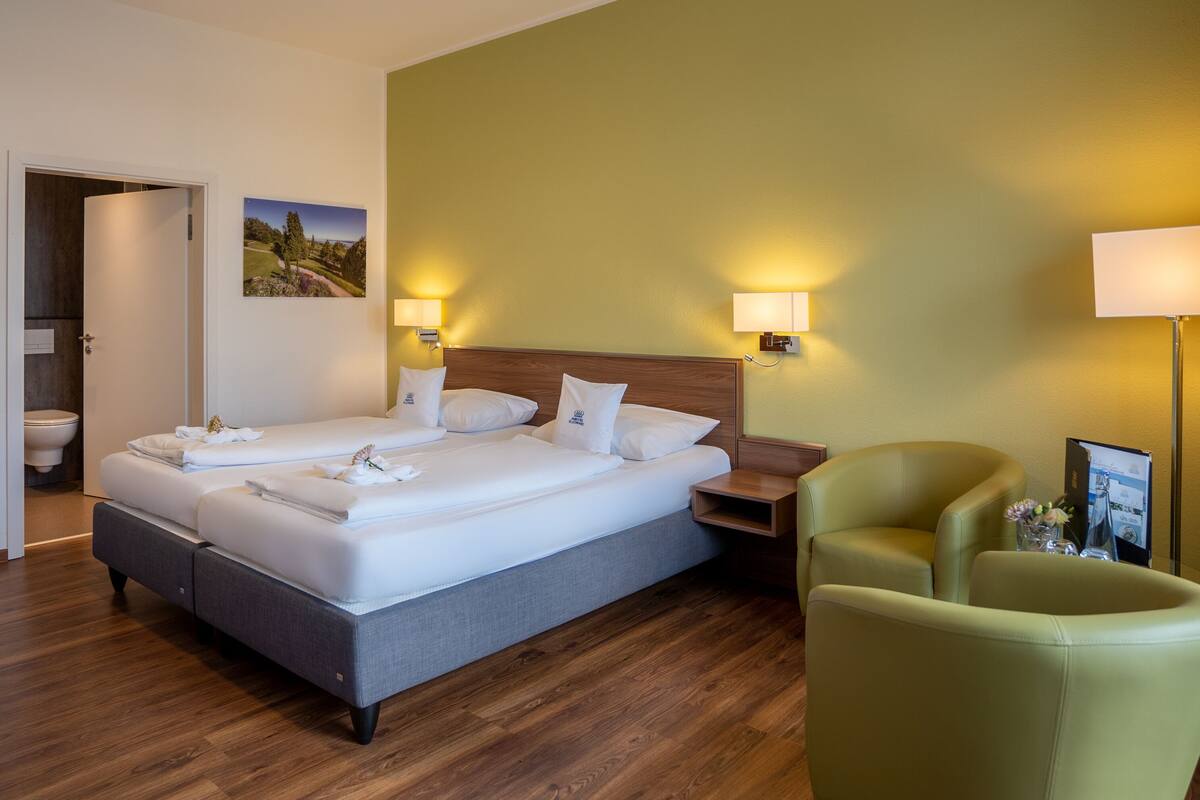 A hotel room with a large bed, two bedside tables, a green armchair, and a wooden floor.