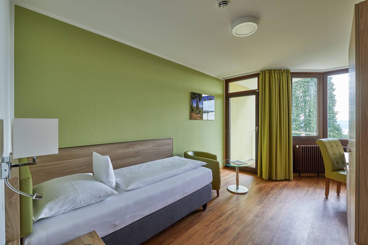 A hotel room with a bed, a chair, a small table, and a window with curtains.