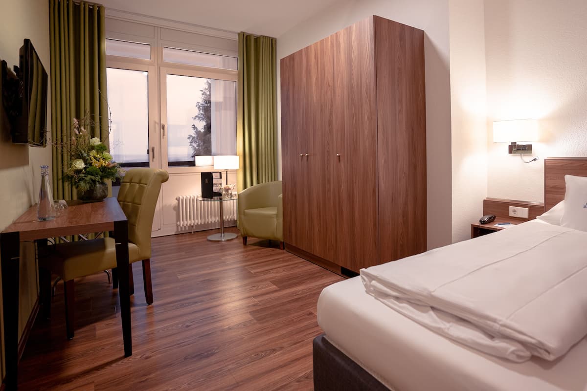A hotel room with a bed, a desk with chairs, a wardrobe, a lamp, and a view of the outdoors through the window.