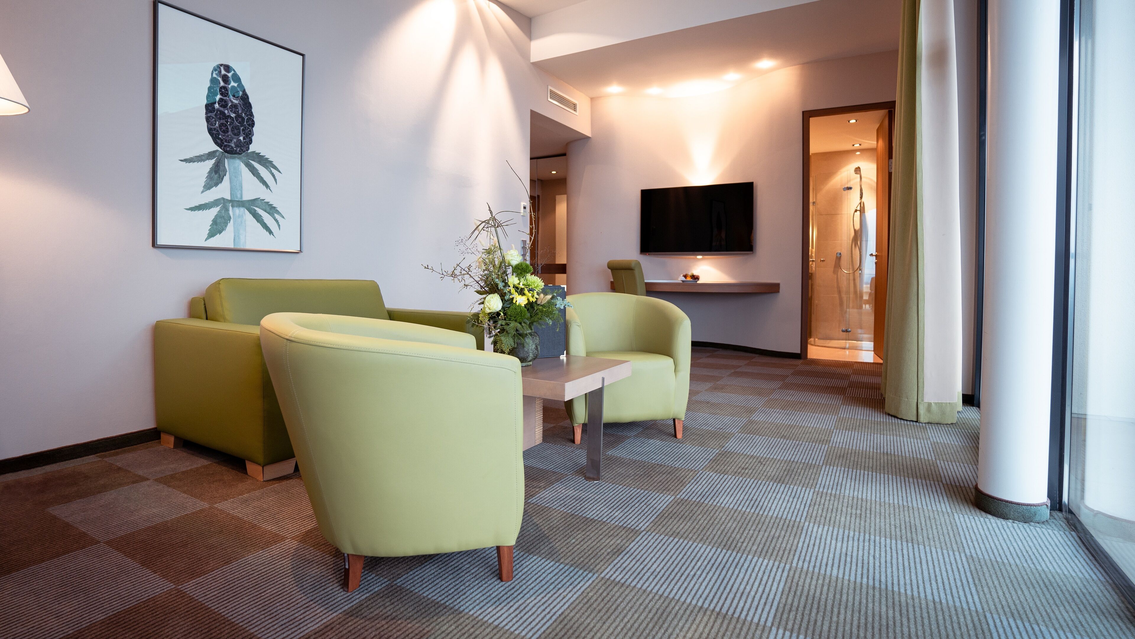 A hotel lobby with a checkered carpet, two yellow armchairs, a small table with a vase, a framed artwork on the wall, and a television.
