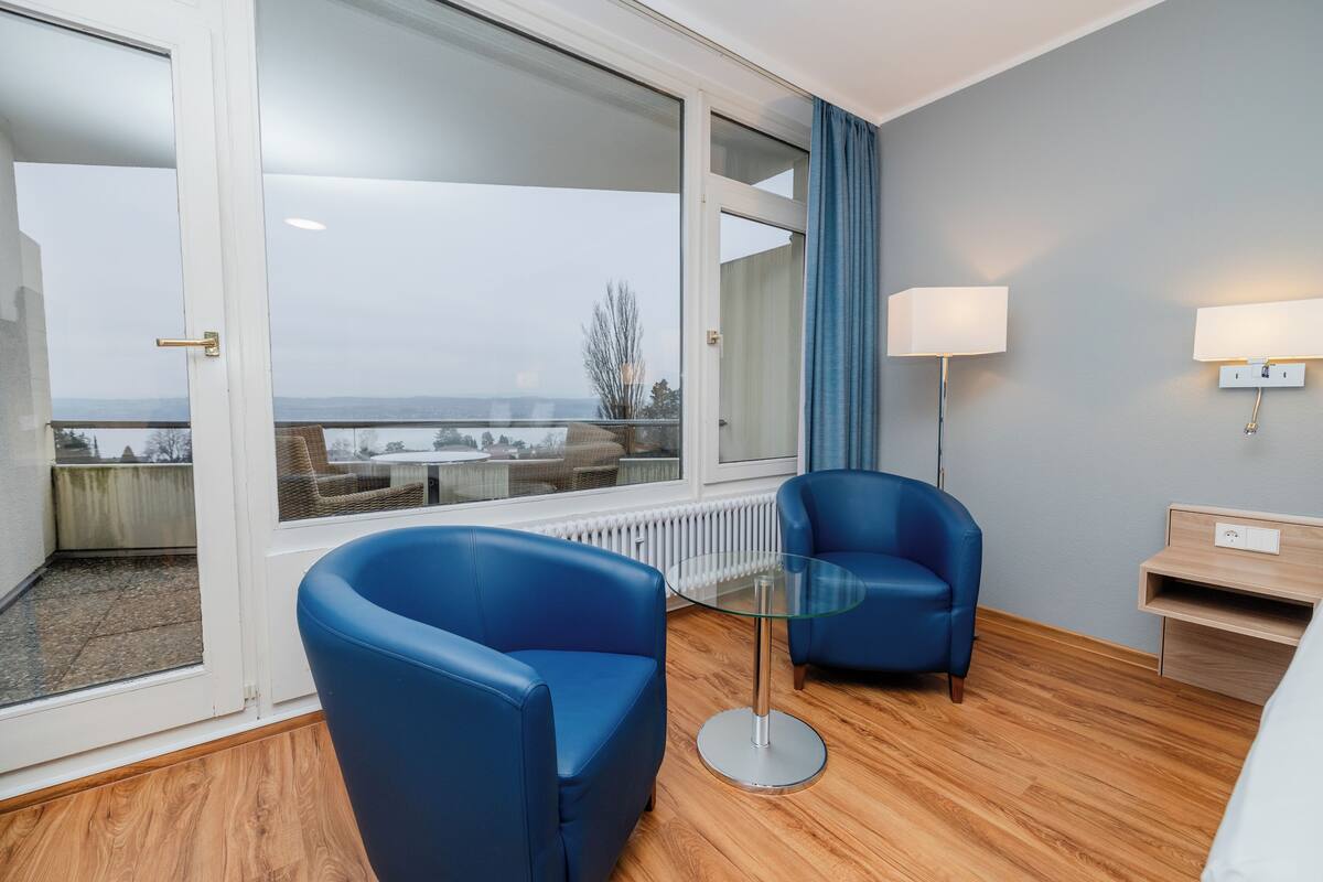 A modern hotel room with a large window, two blue armchairs, a glass table, a lamp, and a wooden floor.