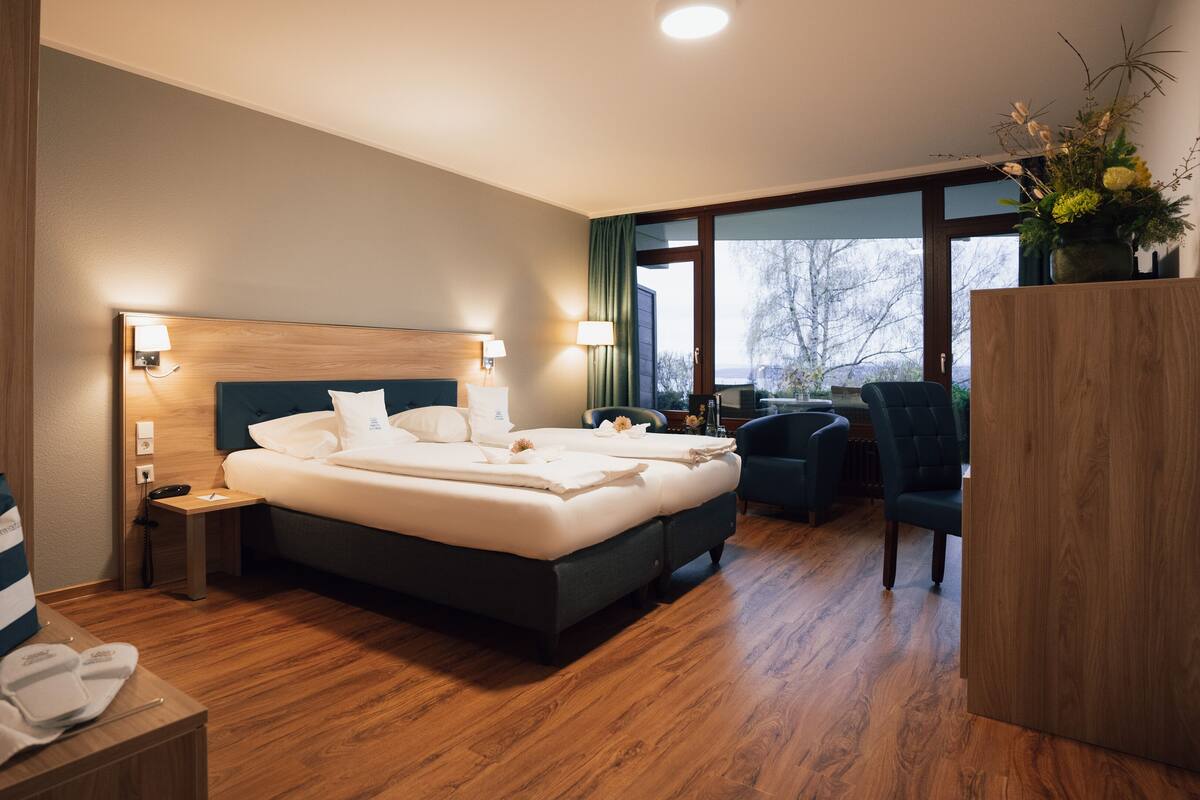 A modern hotel room with a large bed, bedside lamps, a wooden nightstand, a desk with a chair, and a view of the outdoors through the window.