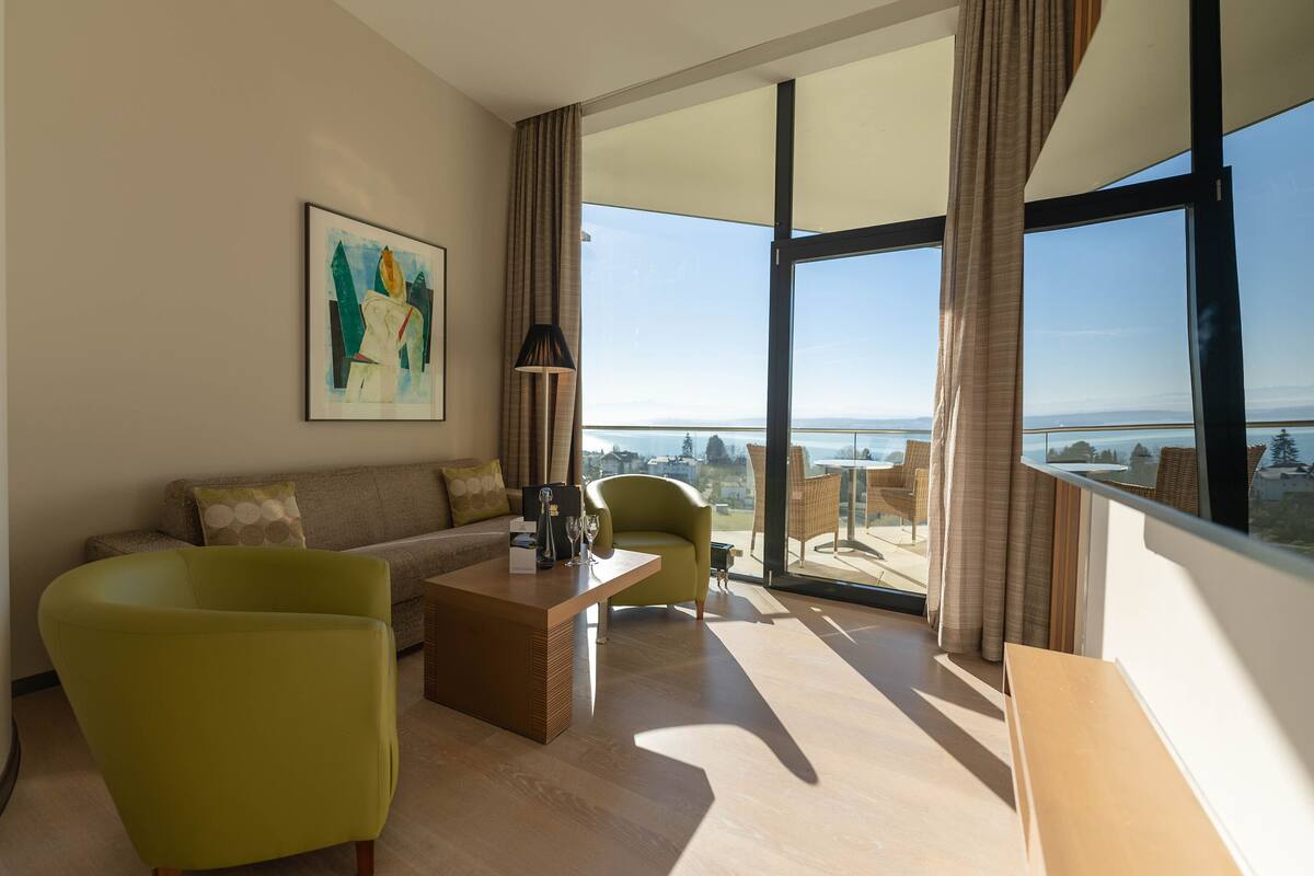 A modern hotel room with a large balcony, a sofa, a coffee table, and a painting on the wall.