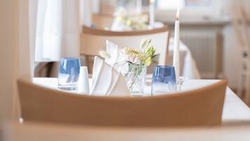 A dining table with a candle, glasses, and a vase with flowers, set in a room with a window and curtains.