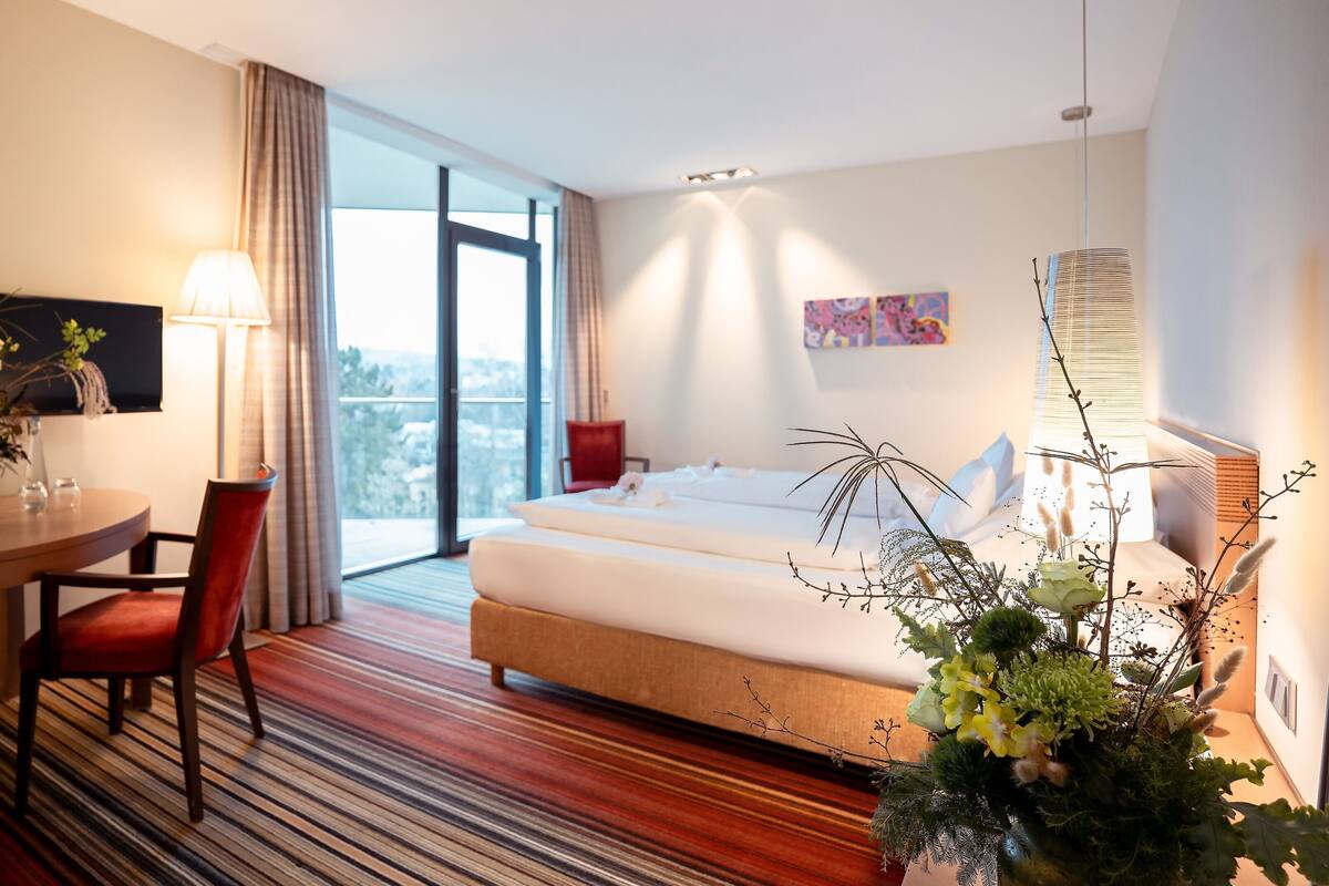 A hotel room with a bed, a desk with a chair, a window with a view, and a plant.