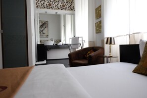 Una habitación de hotel con una cama, una silla, una mesita de noche con lámpara, un baño visible a través de una puerta abierta y obras de arte en las paredes.