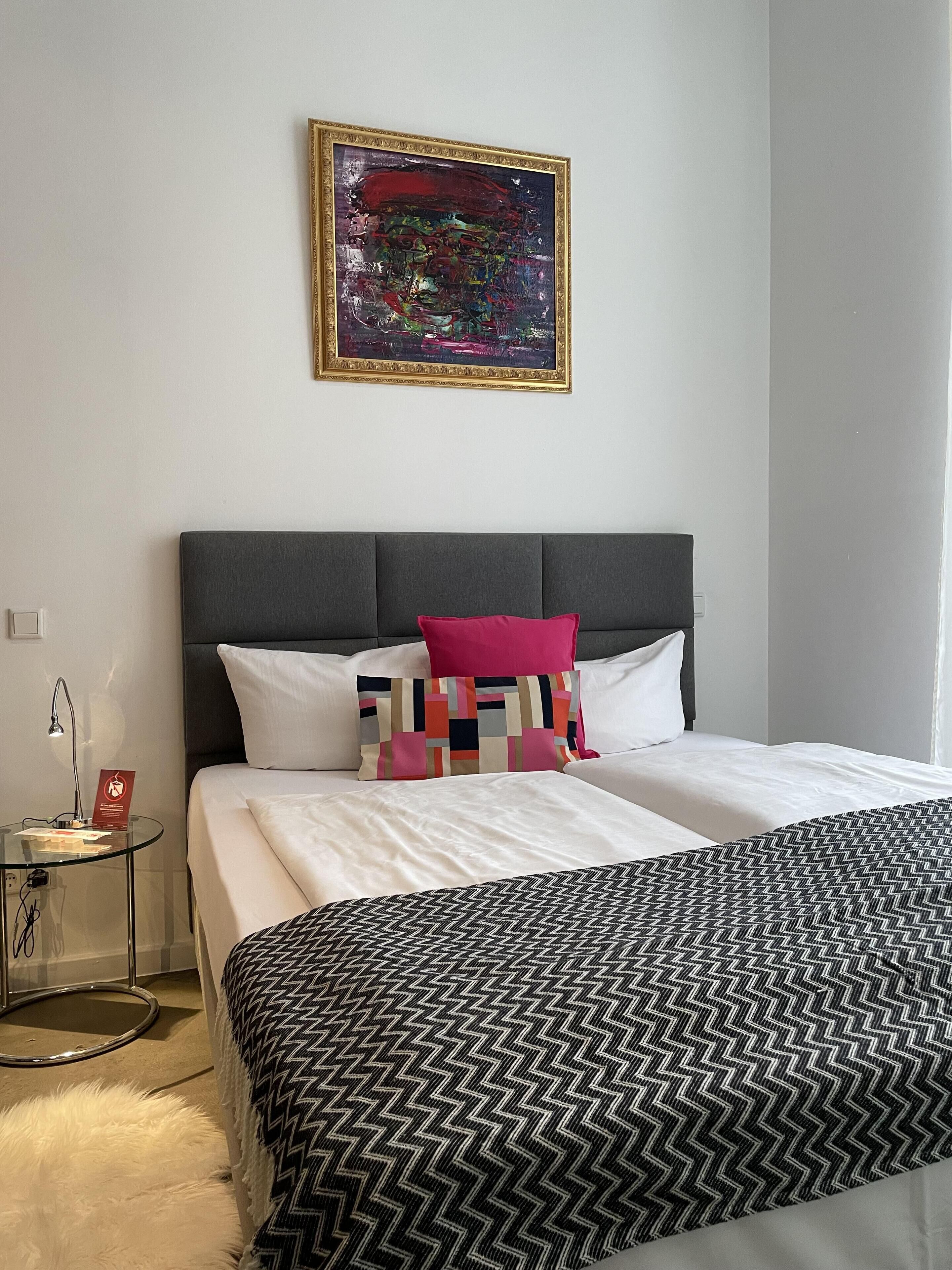 Una habitación de hotel con una cama grande, una mesita auxiliar pequeña, una lámpara, un cuadro abstracto enmarcado y una alfombra blanca y esponjosa.