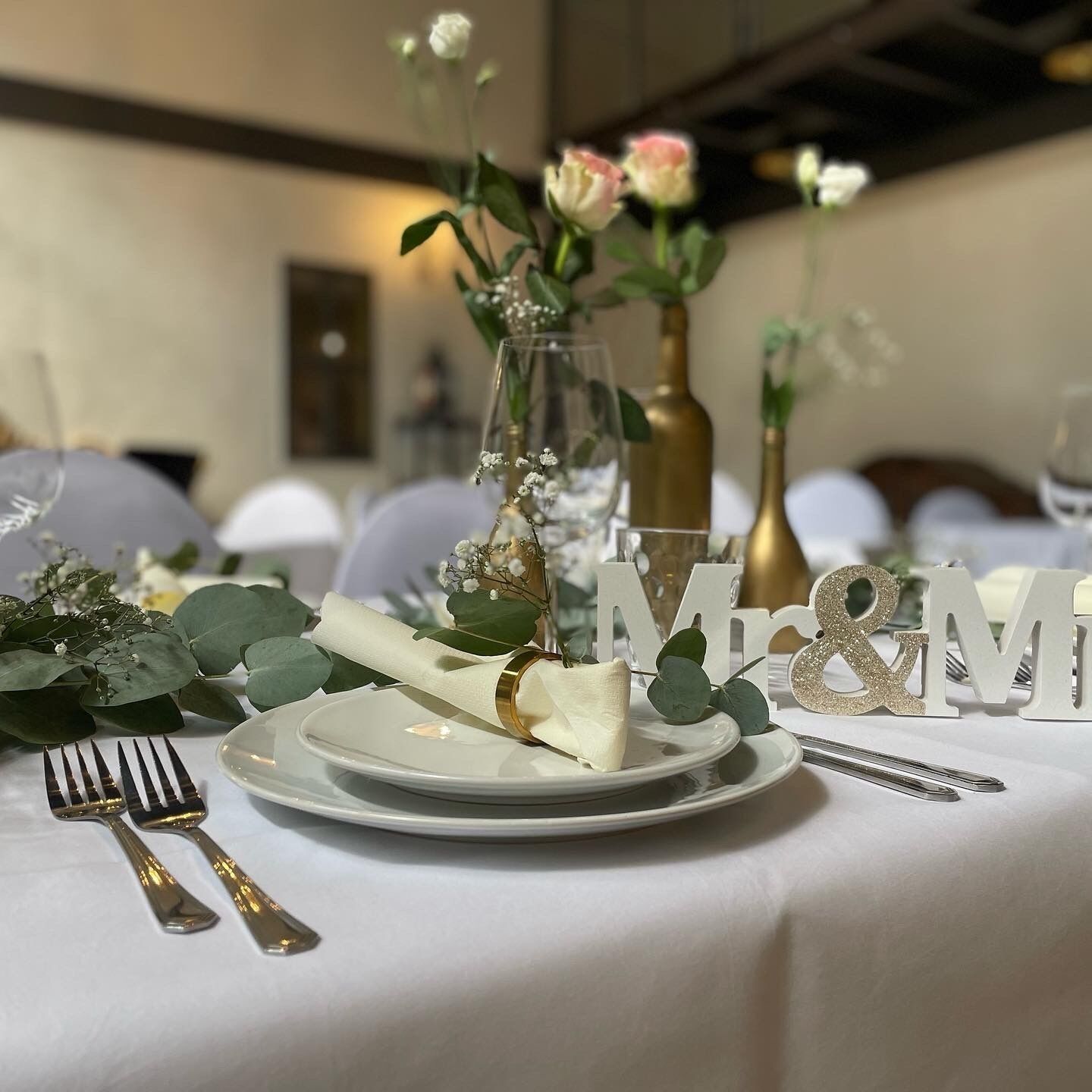 A table set with a white plate, a gold fork, a gold knife, and a gold spoon. There is a glass vase with flowers and a gold bottle in the background.