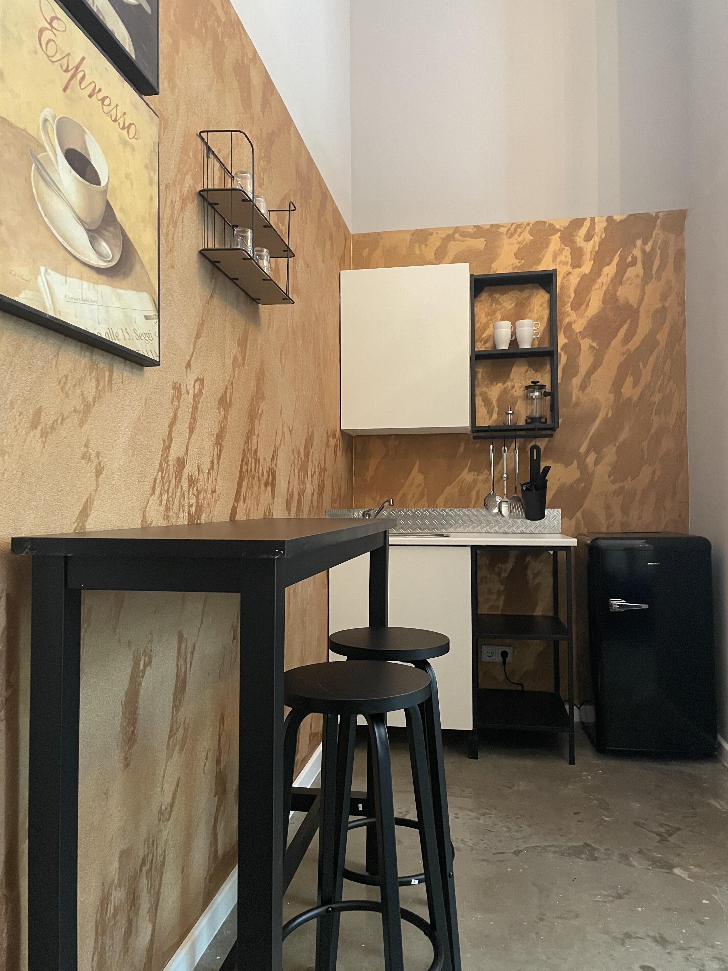Une cuisine compacte avec un plan de travail blanc, un réfrigérateur noir et une machine à café. On y trouve deux tabourets noirs et une petite étagère avec des tasses et une cafetière.