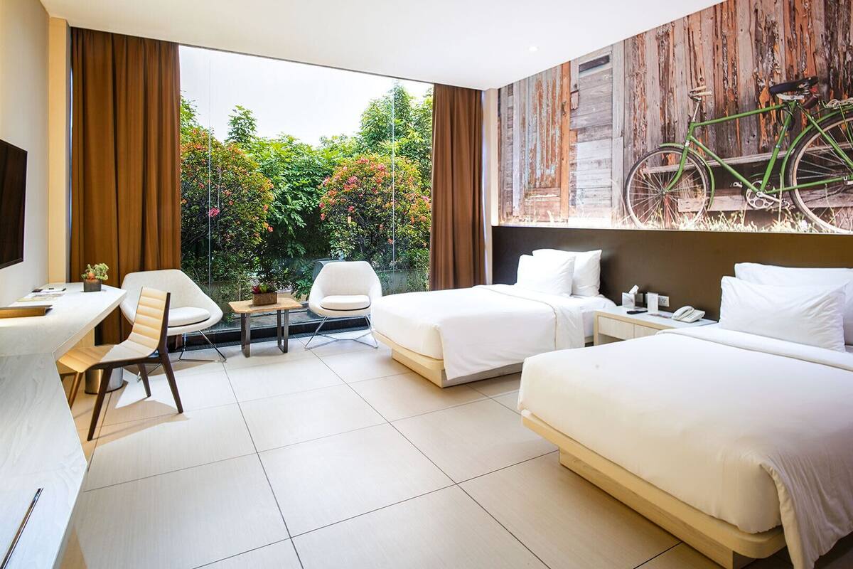 A hotel room with two beds, a desk, a chair, a TV, and a view of greenery and a bicycle outside.