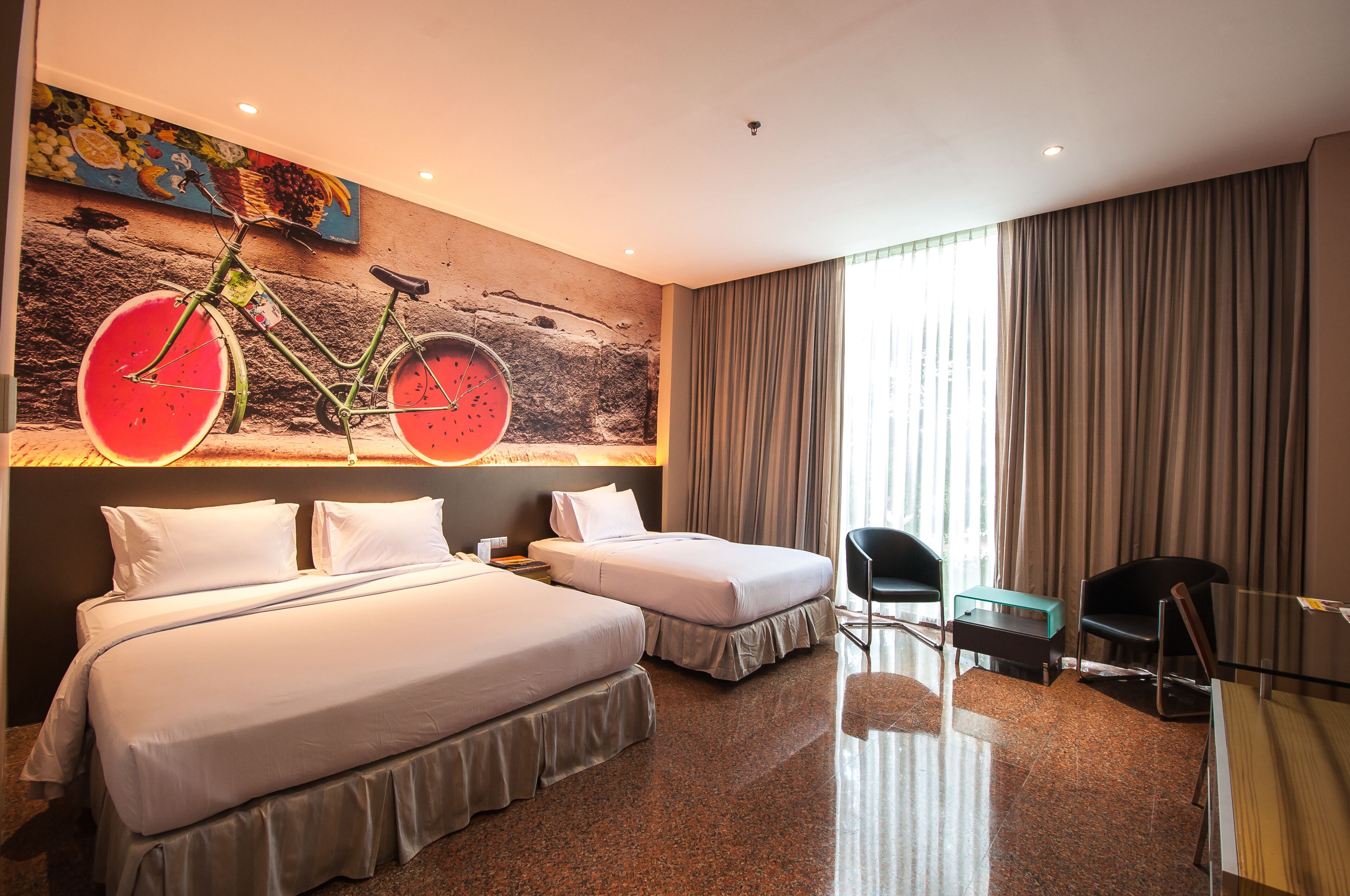 A hotel room with two beds, a bicycle mural, a window with curtains, and a small table.