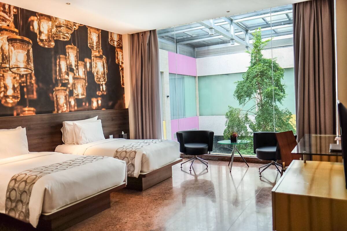 A hotel room with two beds, a glass table with chairs, a TV, and a large window with a view of greenery.