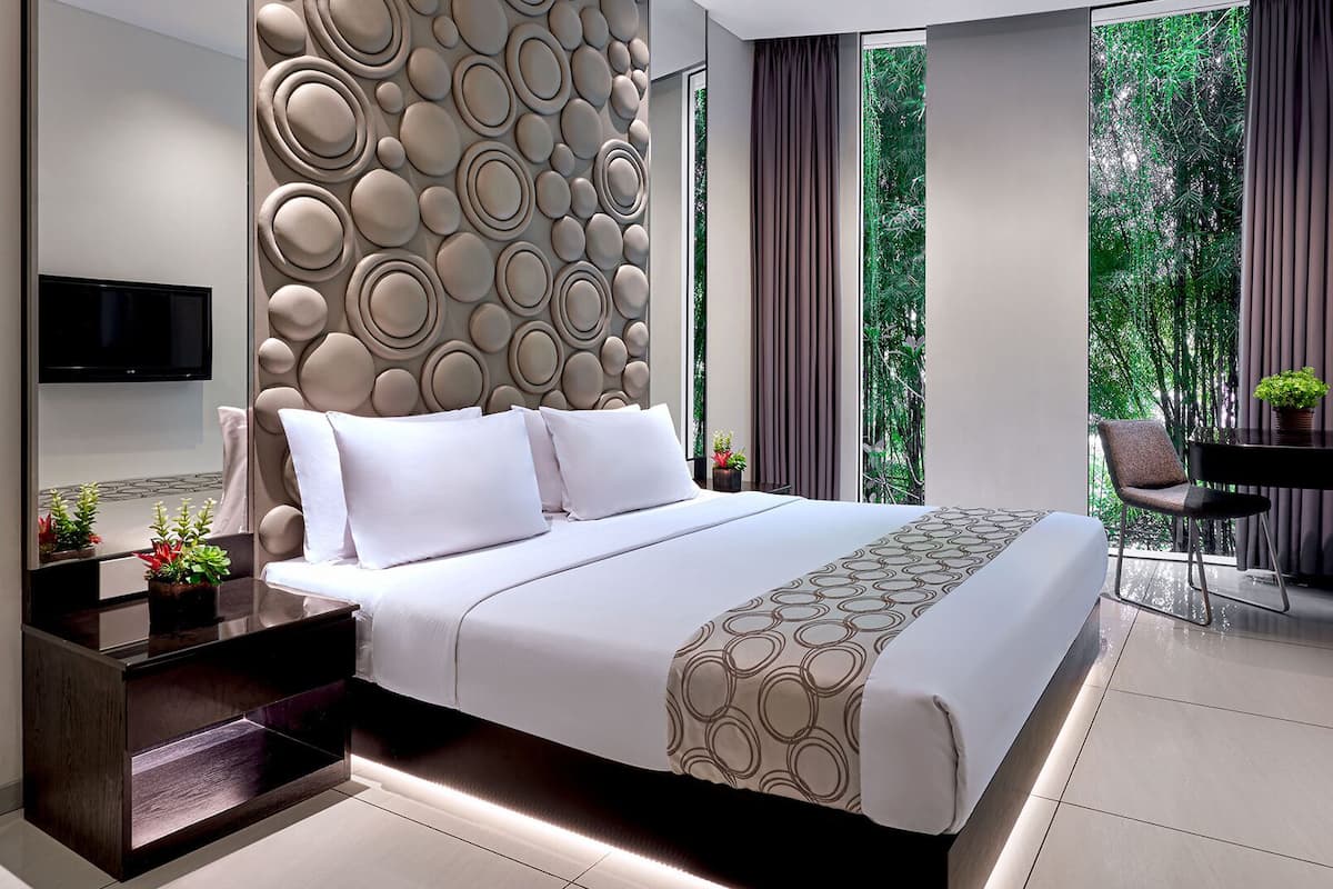 A modern bedroom with a large bed, a TV, a desk, and a view of greenery.