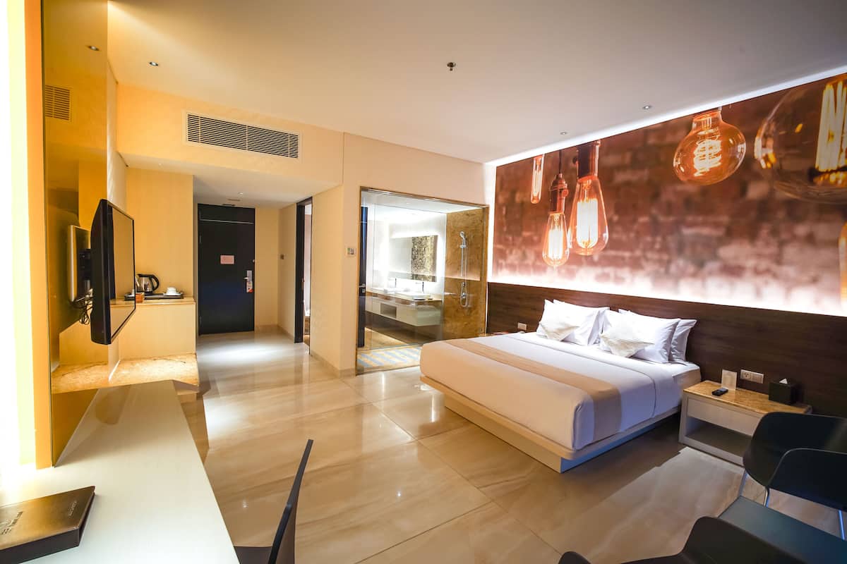 A modern hotel room with a bed, bedside tables, a TV, and a bathroom visible through an open door.