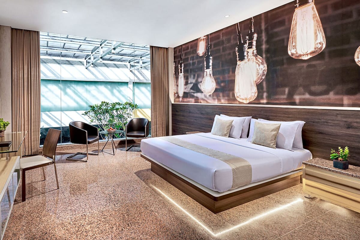 A modern hotel room with a large bed, a flat-screen TV, a desk, and a seating area with a view of greenery.