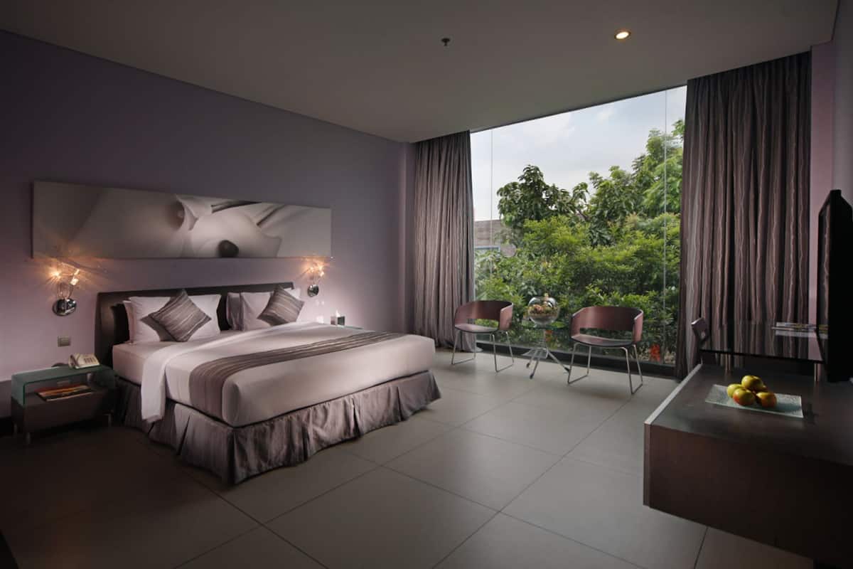 A modern bedroom with a large bed, a nightstand, a chair, a small table with fruit, and a view of greenery outside.