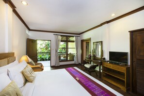 A hotel room with a large bed, a desk with a television, and a view of greenery outside. - Adi Cottage (Ubud)