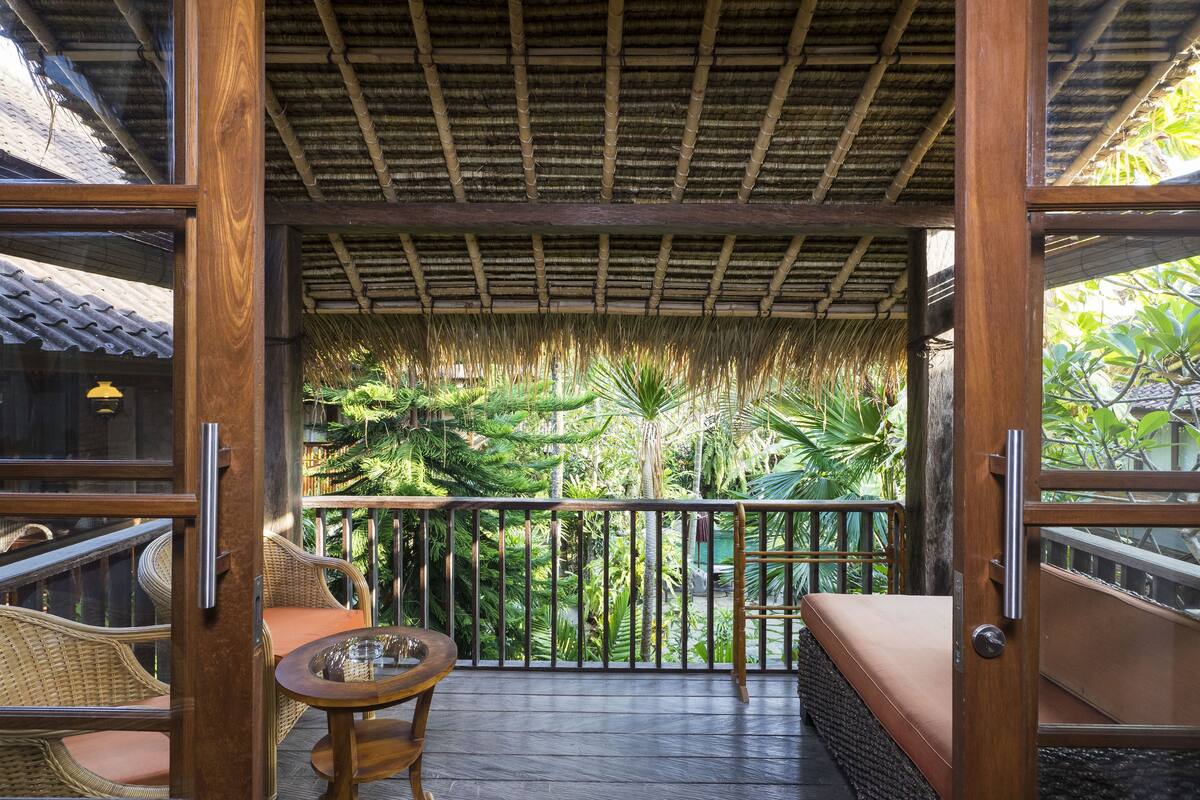 A wooden balcony with a thatched roof, wicker furniture, and a view of lush greenery.