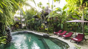 A pool with red lounge chairs and a thatched-roof structure in a lush garden. - Adi Cottage (Ubud)