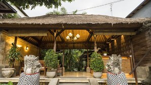 A traditional wooden structure with a thatched roof, stone steps leading up to a covered entrance, and two stone lion statues flanking the entrance. - Adi Cottage (Ubud)