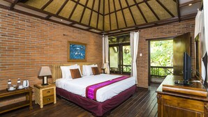 A bedroom with a large bed, wooden nightstands, a TV, and a view of greenery outside. - Adi Cottage (Ubud)