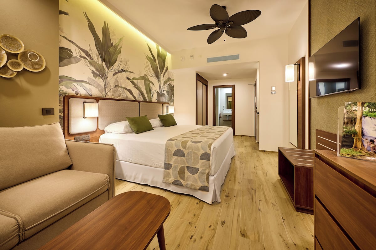 A hotel room with a bed, a sofa, a wooden table, a ceiling fan, a TV, and a wall with a plant mural.