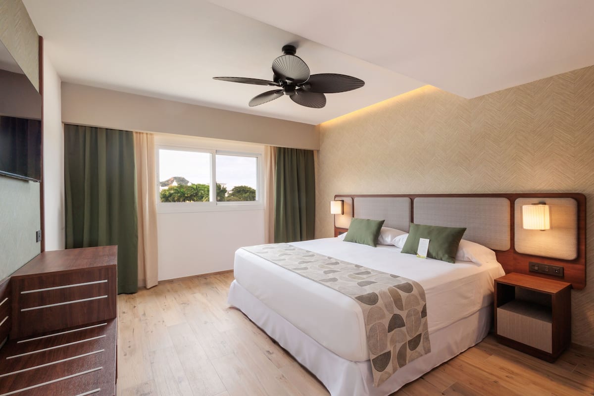 A hotel room with a bed, a ceiling fan, a nightstand with a lamp, a wooden dresser, and a window with curtains.