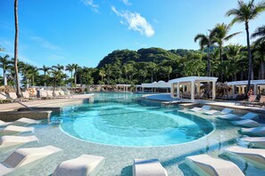 A large, clear swimming pool with multiple lounge areas and palm trees.