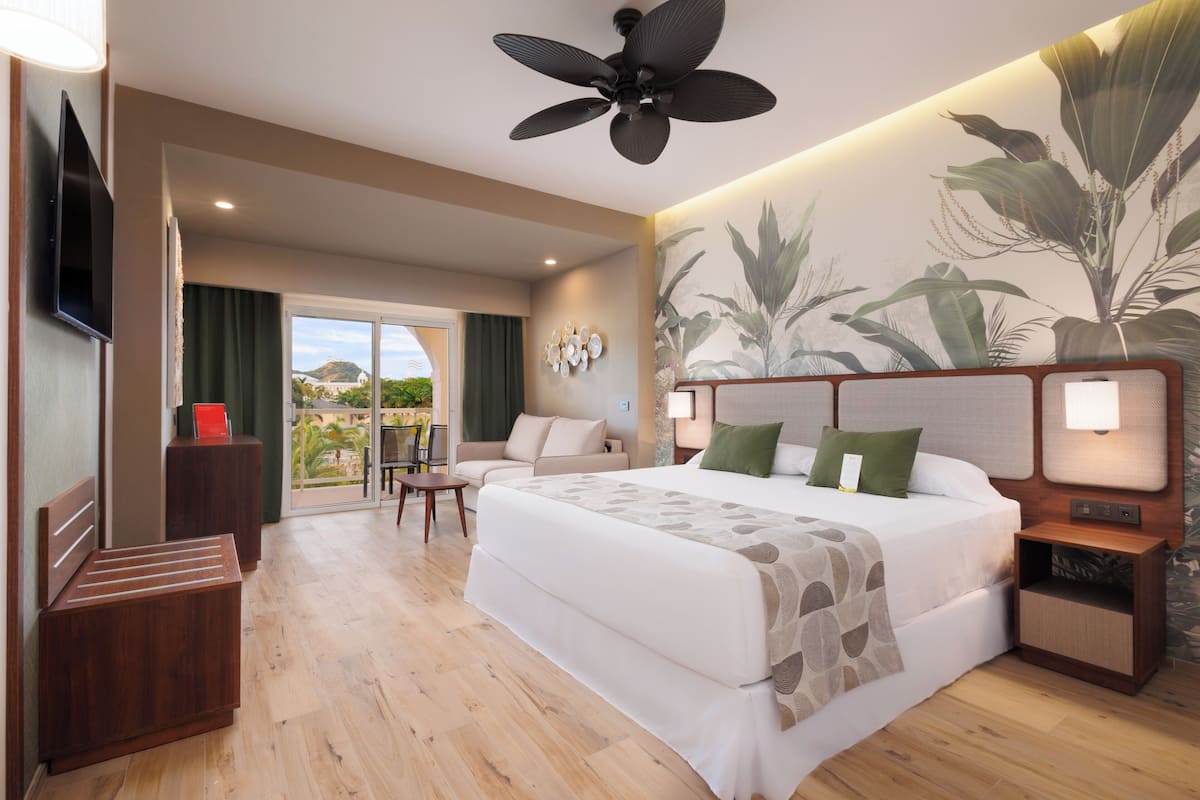 A hotel room with a large bed, a ceiling fan, a TV, a small table, and a view of the outdoors.