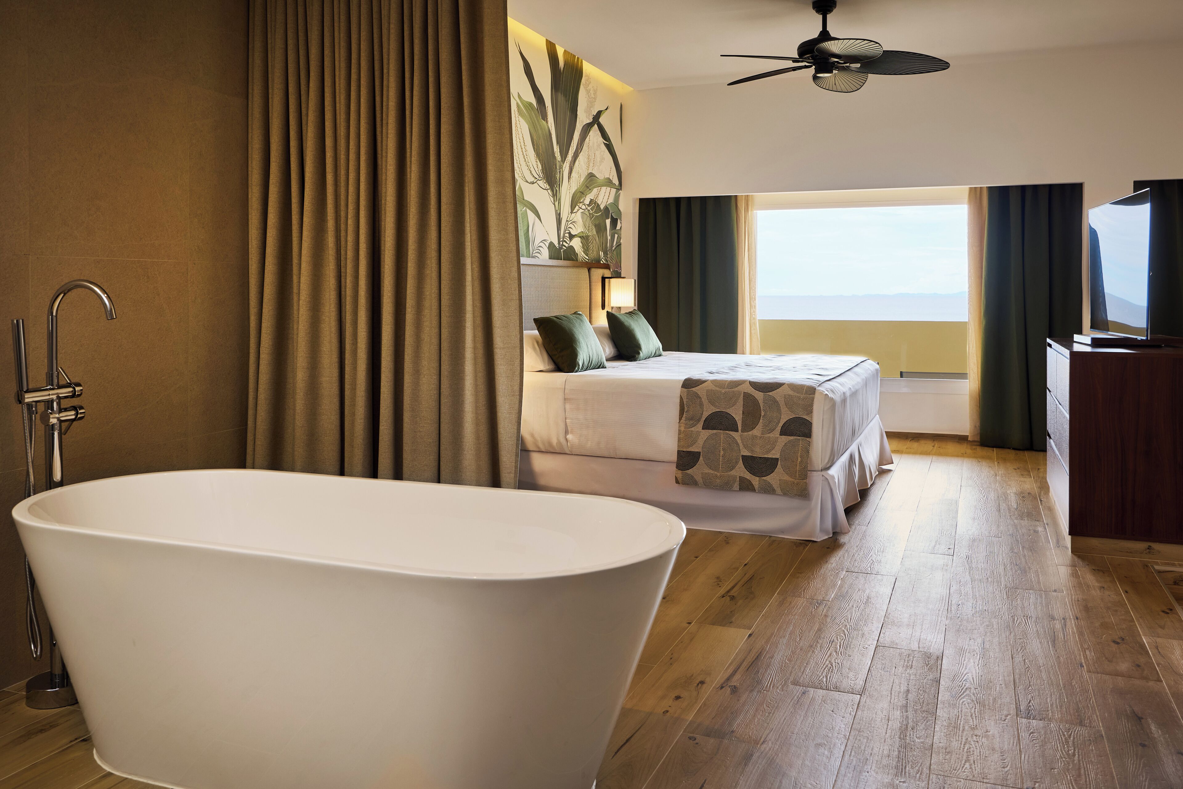 Ein modernes Hotelzimmer mit einer großzügigen Badewanne, einem Bett mit grünen Kissen, einem Fernseher und Blick aufs Meer.