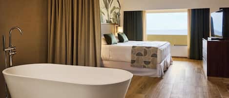 A modern hotel room with a large bathtub, a bed with green pillows, a television, and a view of the sea.
