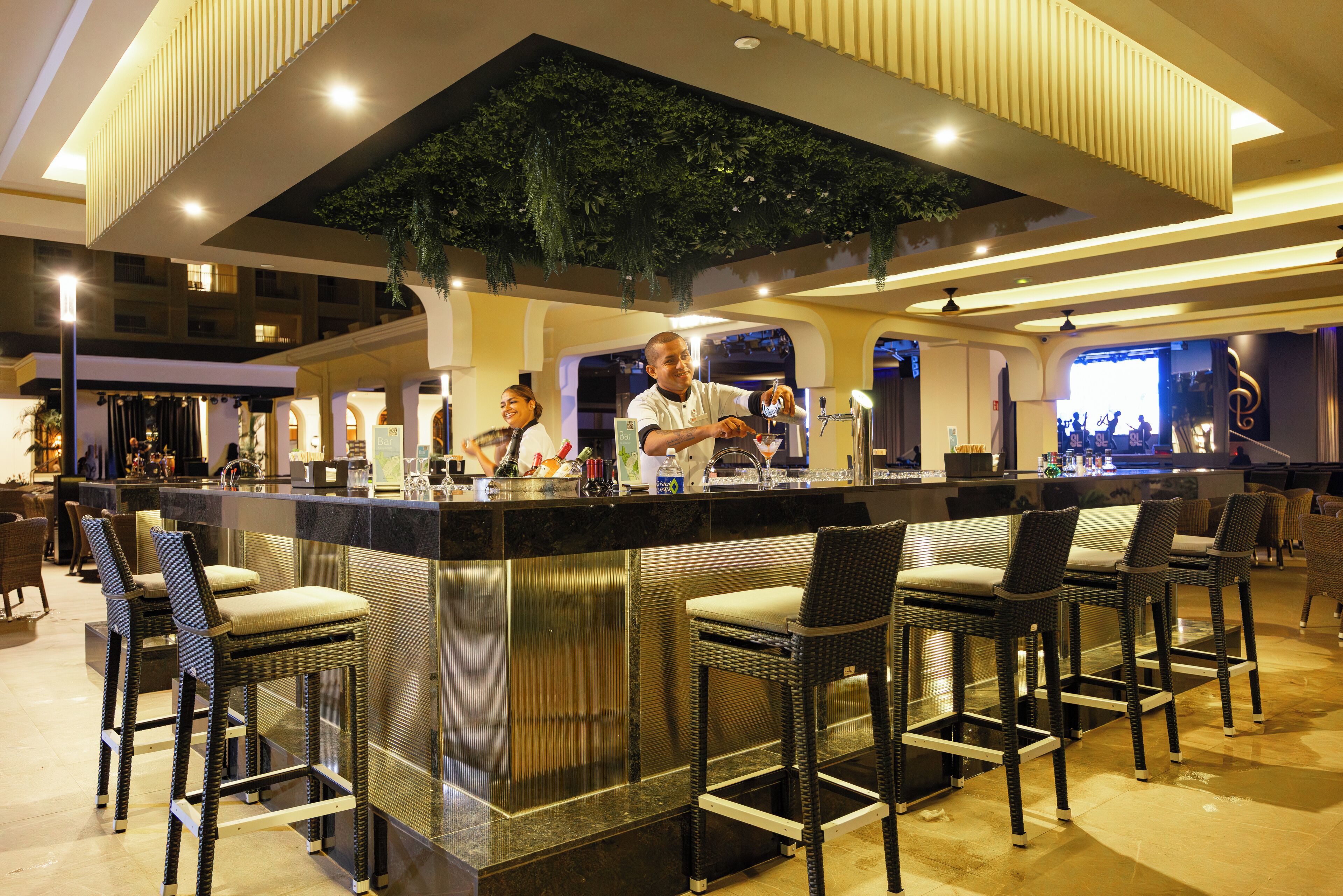 A bar area with a central counter, bar stools, and a ceiling with greenery.